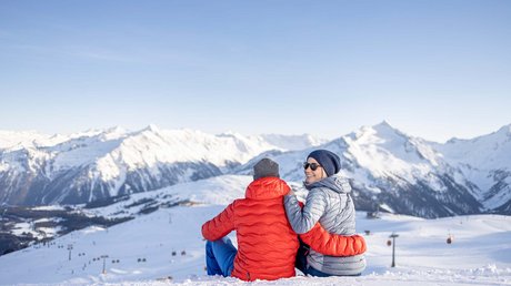 Pictures & Impressions • Der Grubacher: Hotel in Gerlos Couple sitting in the snow enjoying the view of snow-covered Alps under a blue sky