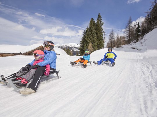 Erlebnisse im Zillertal • Der Grubacher: Biohotel in Gerlos Familie rodelt lachend auf verschneiter Piste mit Winterlandschaft im Hintergrund