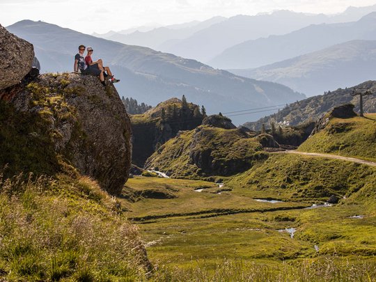 Erlebnisse im Zillertal • Der Grubacher: Biohotel in Gerlos Paar sitzt auf einem großen Felsen mit Blick auf Wiesen, Bäche und Berge