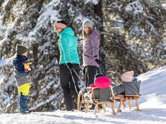 Erlebnisse im Zillertal • Der Grubacher: Biohotel in Gerlos Zwei Frauen ziehen Kinder auf Schlitten durch verschneiten Winterwald